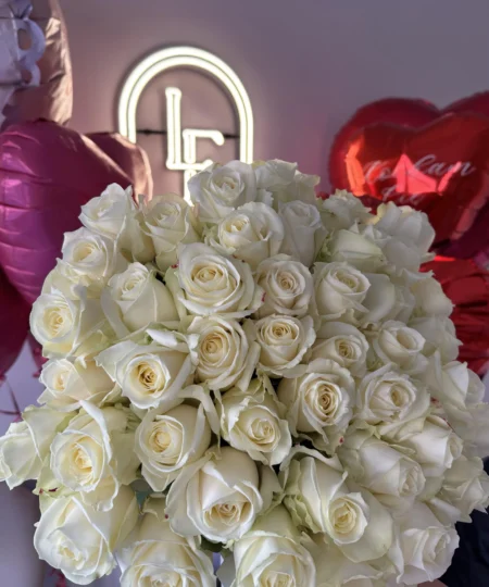 Duży bukiet białych róż z długimi łodygami, ułożony na tle balonów w kształcie serca i logo Lu La Fleur