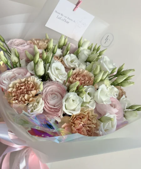 Widok z boku bukietu White Tenderness z brzoskwiniowymi goździkami, białą eustomą i jasnoróżowymi jaskrami w opalizującym papierze