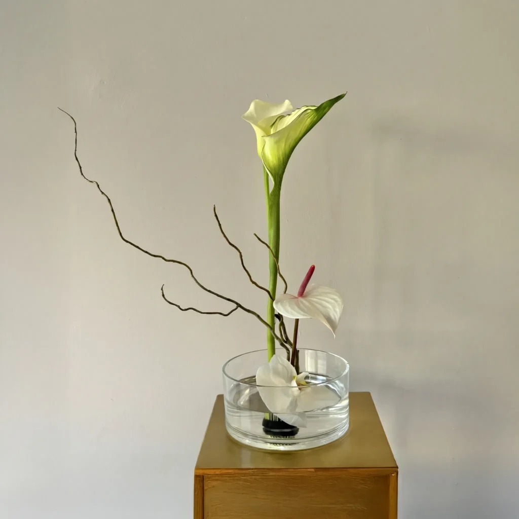 Minimalistyczna kompozycja ikebana z kallą i anthurium w szklanej misie, ułożona na kenzanie w eleganckim stylu.